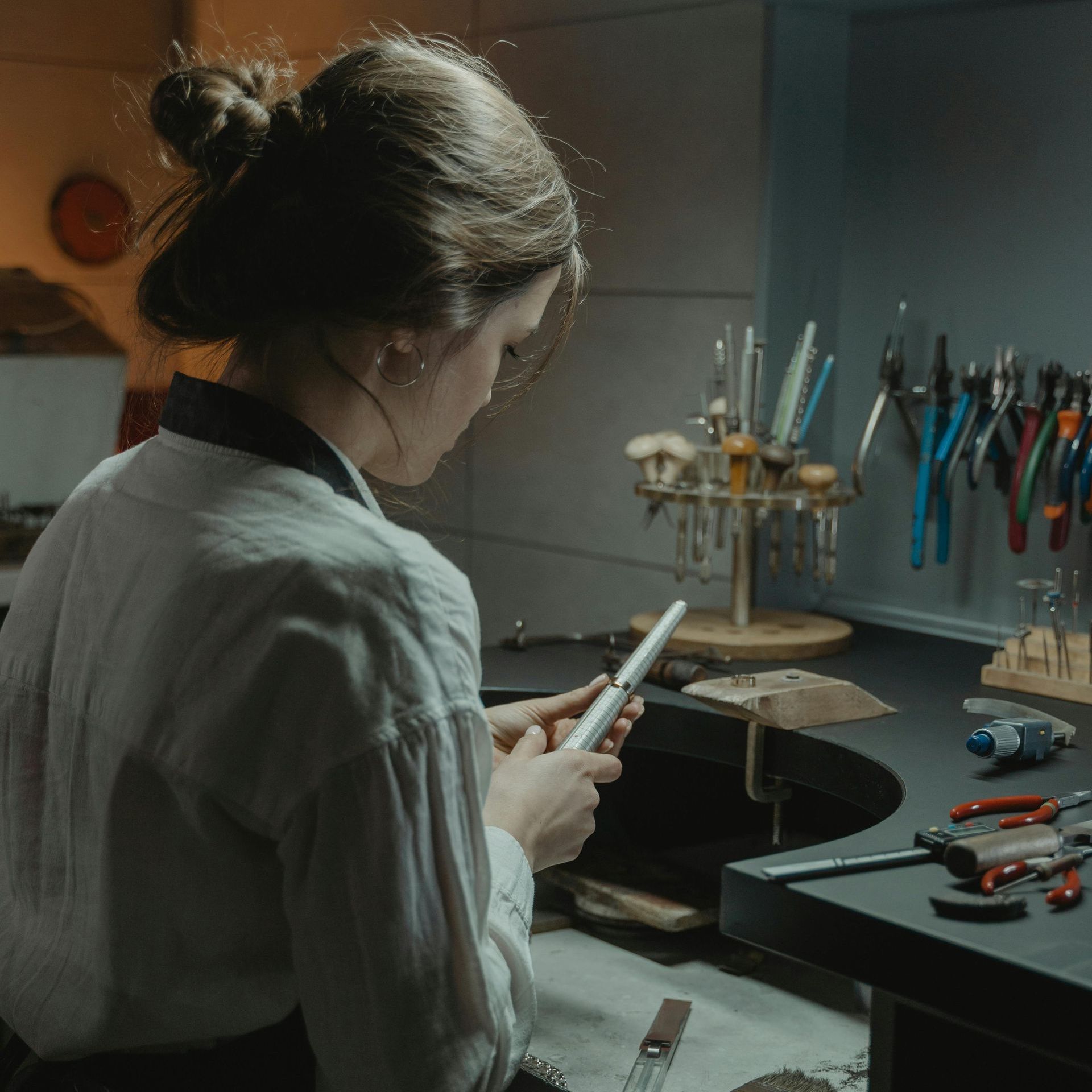 Read Full Letter of Comment Woman working at a jewelry bench, examining a white object. Tools and supplies on the dark workspace.