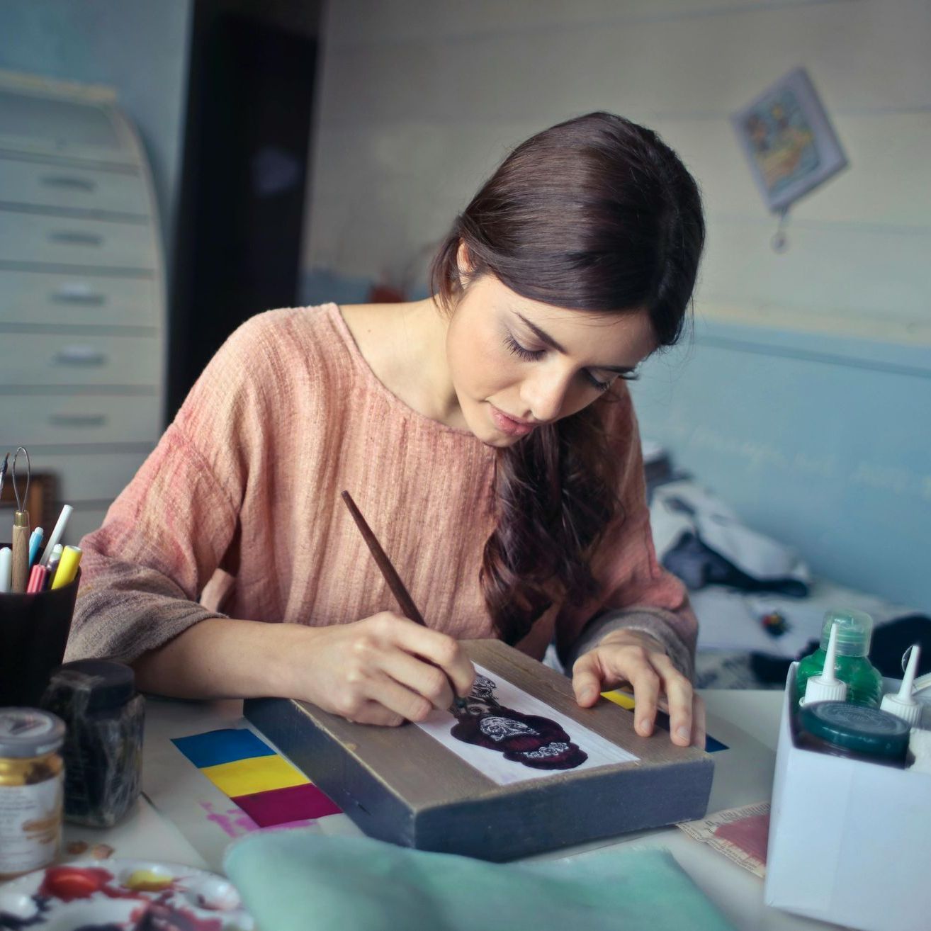 Read Full Letter of Comment Woman painting at a desk, wearing a pink top, holding a paintbrush. Paints and art supplies are scattered around.