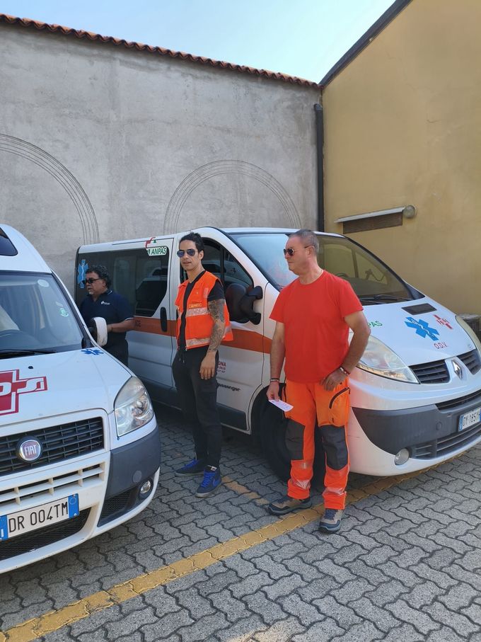 Tre soccorritori della Croce Rossa con giubbotti arancioni parlano accanto a un'ambulanza all'aperto.