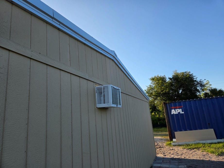 Side of a beige building with a window air conditioner, beside a blue container and trees under a clear sky.