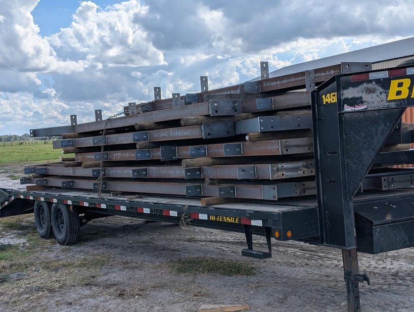 Stacked steel beams loaded on a flatbed trailer in a gravel lot under a cloudy sky