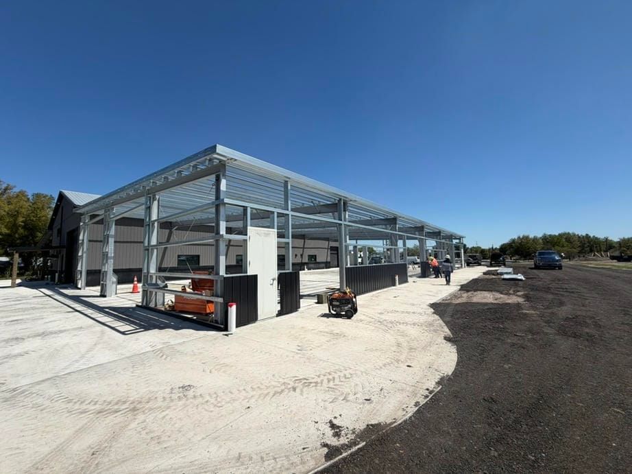 Steel-framed building under construction on a sunny lot, with gravel foreground and parked vehicles nearby