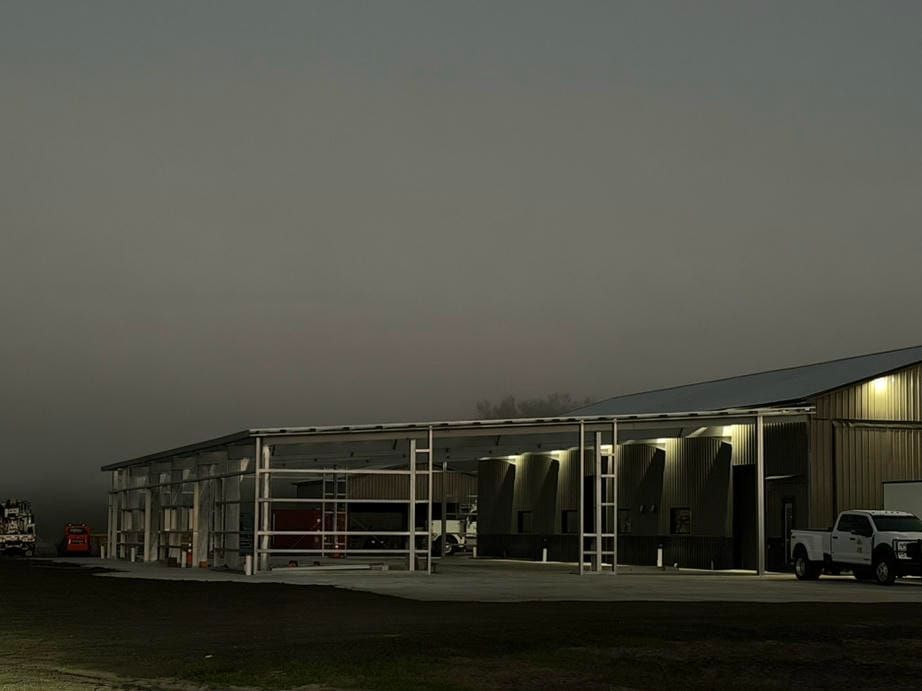 Industrial building at dusk with lit exterior lights and parked vehicles under a gray sky
