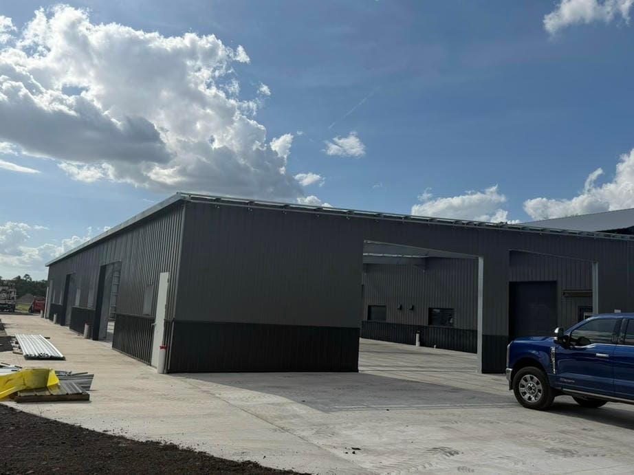Gray industrial building with open bays on a concrete lot under a partly cloudy sky, blue truck at right