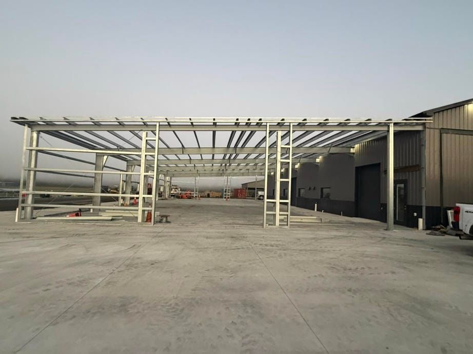Industrial warehouse frame under construction on a concrete lot, with steel beams and a metal-sided building.