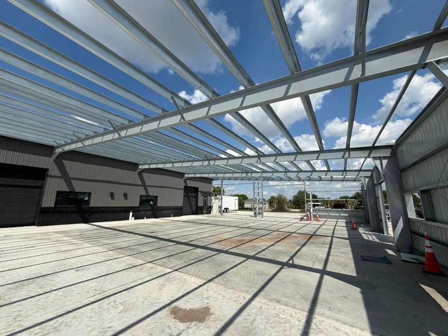 Open steel-framed roof over a concrete lot under construction, with scattered cones and shadows.