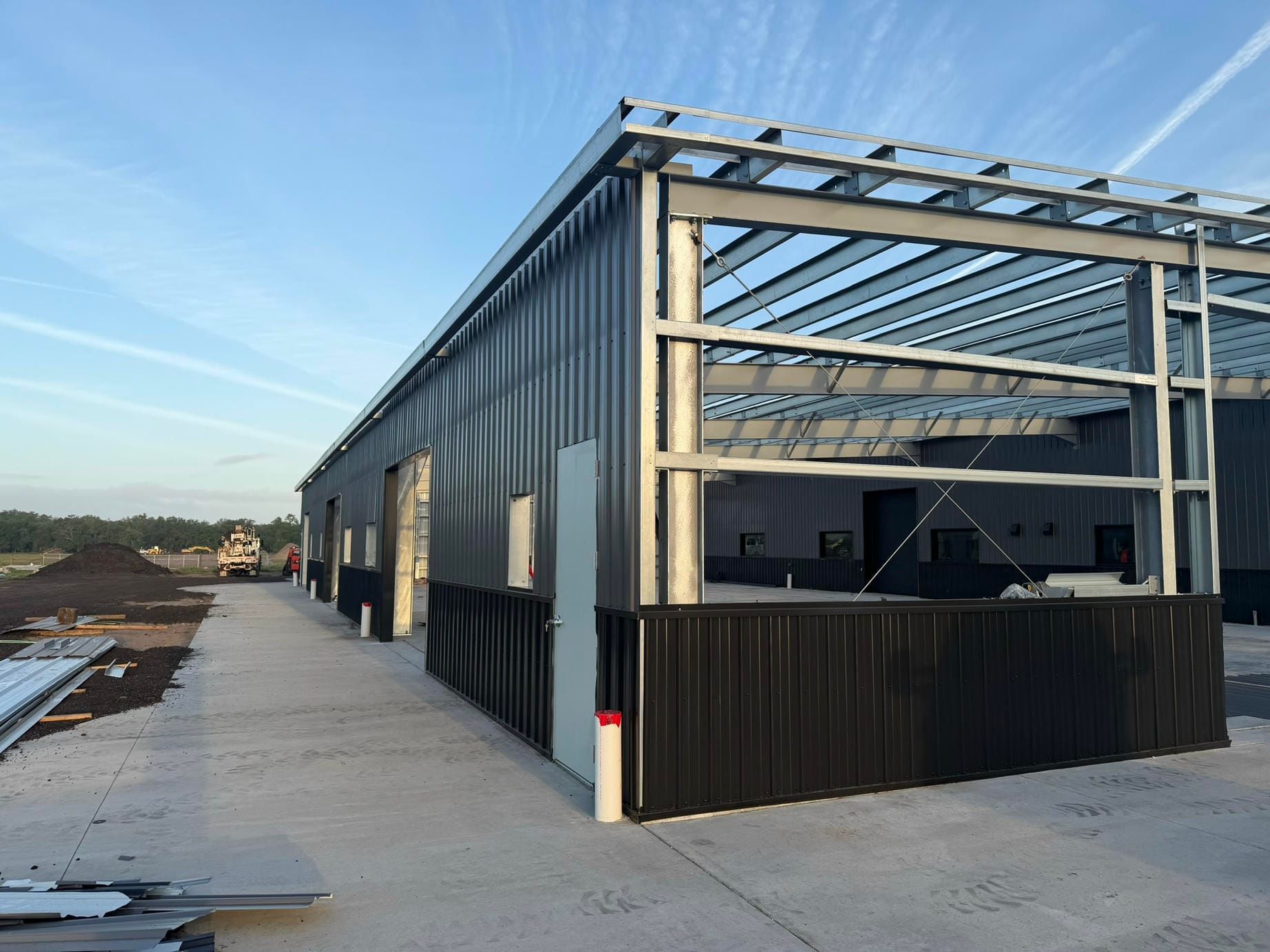 New metal warehouse under construction beside a paved road and open field at sunset