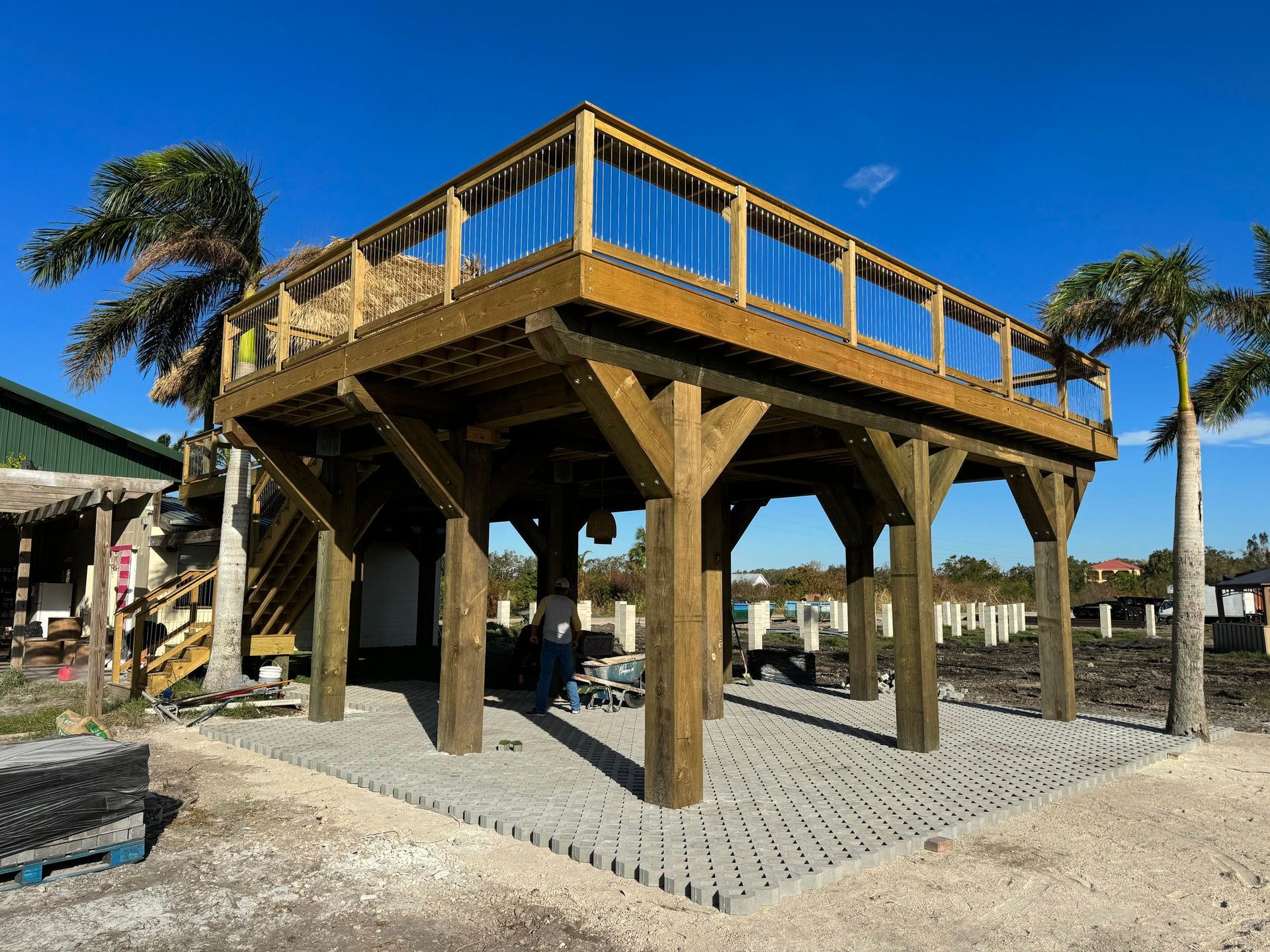 Elevated wooden deck under construction on a gravel lot, with palm trees and blue sky in the background