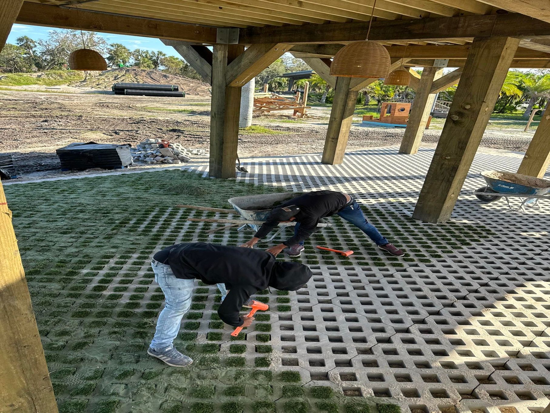 Two people crouch under a wooden shelter on a grated platform, working with orange tools outdoors.