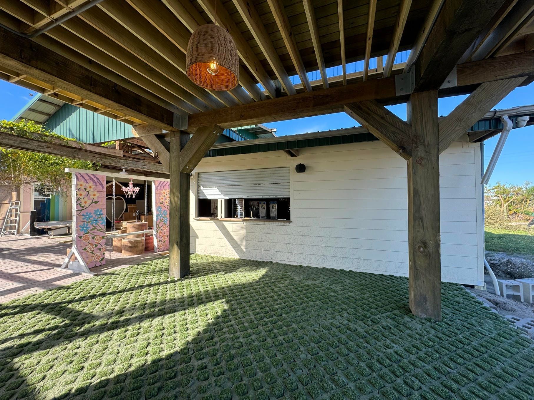 Covered outdoor patio with wooden beams, grass-pattern flooring, and a white building wall in sunlight