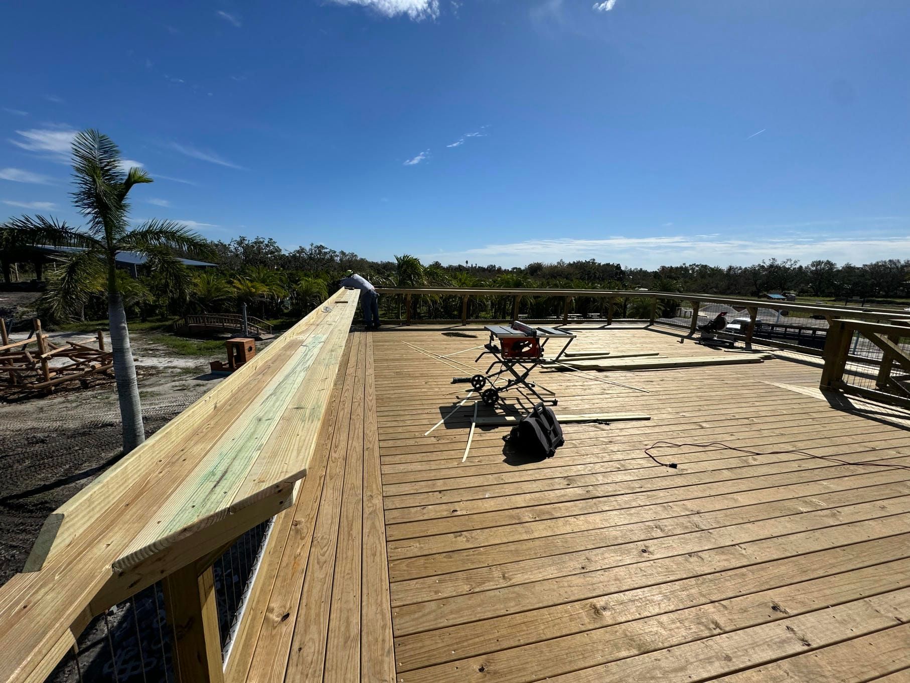 Wooden deck under construction with railings, tools, and a bright blue sky beyond the trees