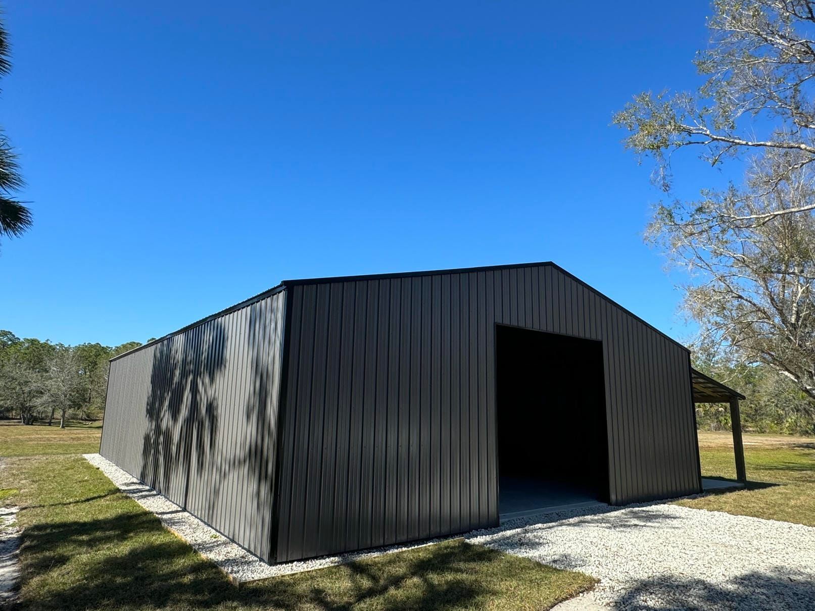 Large dark metal barn-like shed with open doorway on gravel under a blue sky