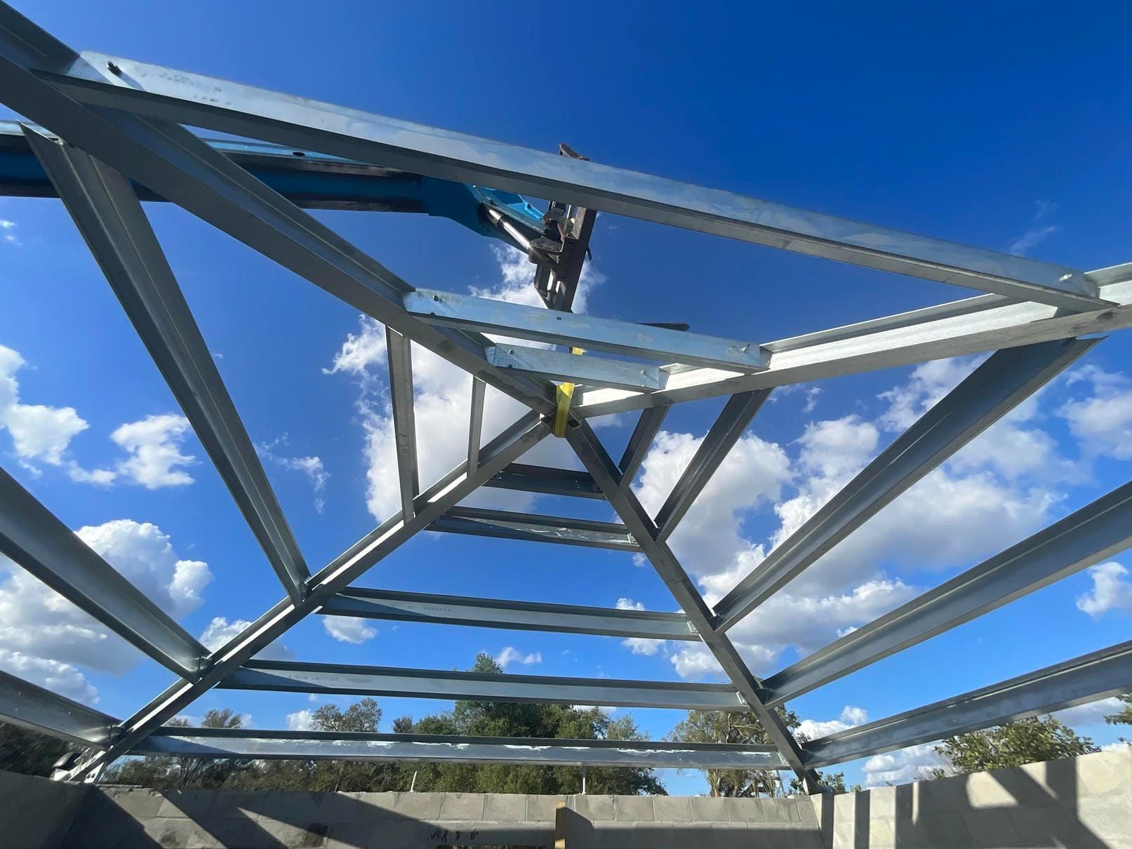 Metal roof truss framework against a bright blue sky with white clouds