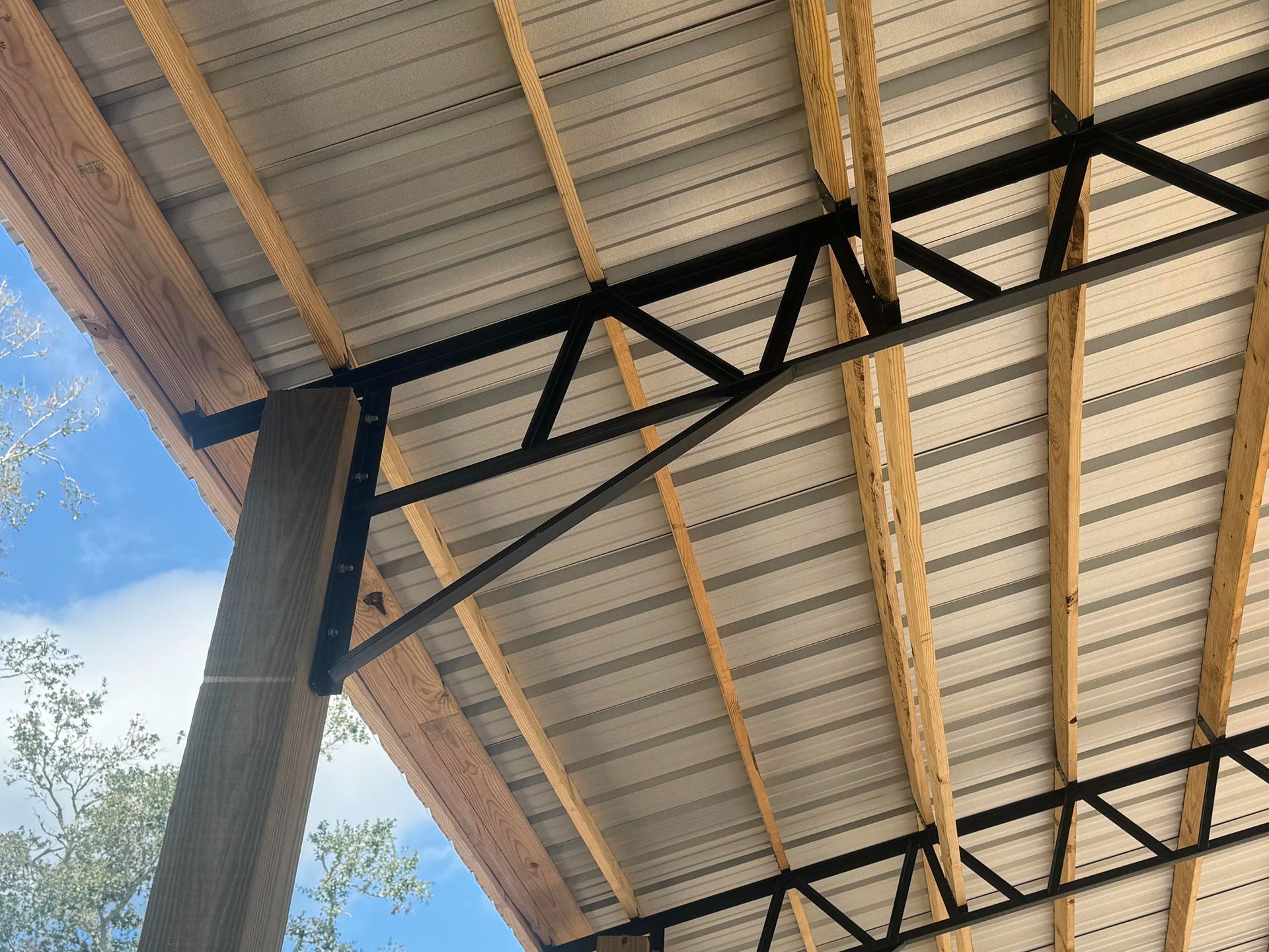 Covered outdoor roof with black steel trusses and light corrugated panels against a blue sky