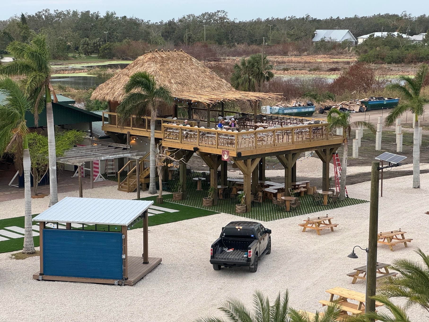 Outdoor resort area with a thatched-roof deck, palm trees, a parked car, and a blue kiosk.