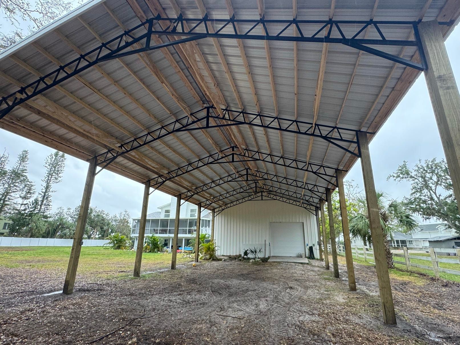 Open-sided metal shed with a curved roof on a gravel lot, surrounded by trees and fields.