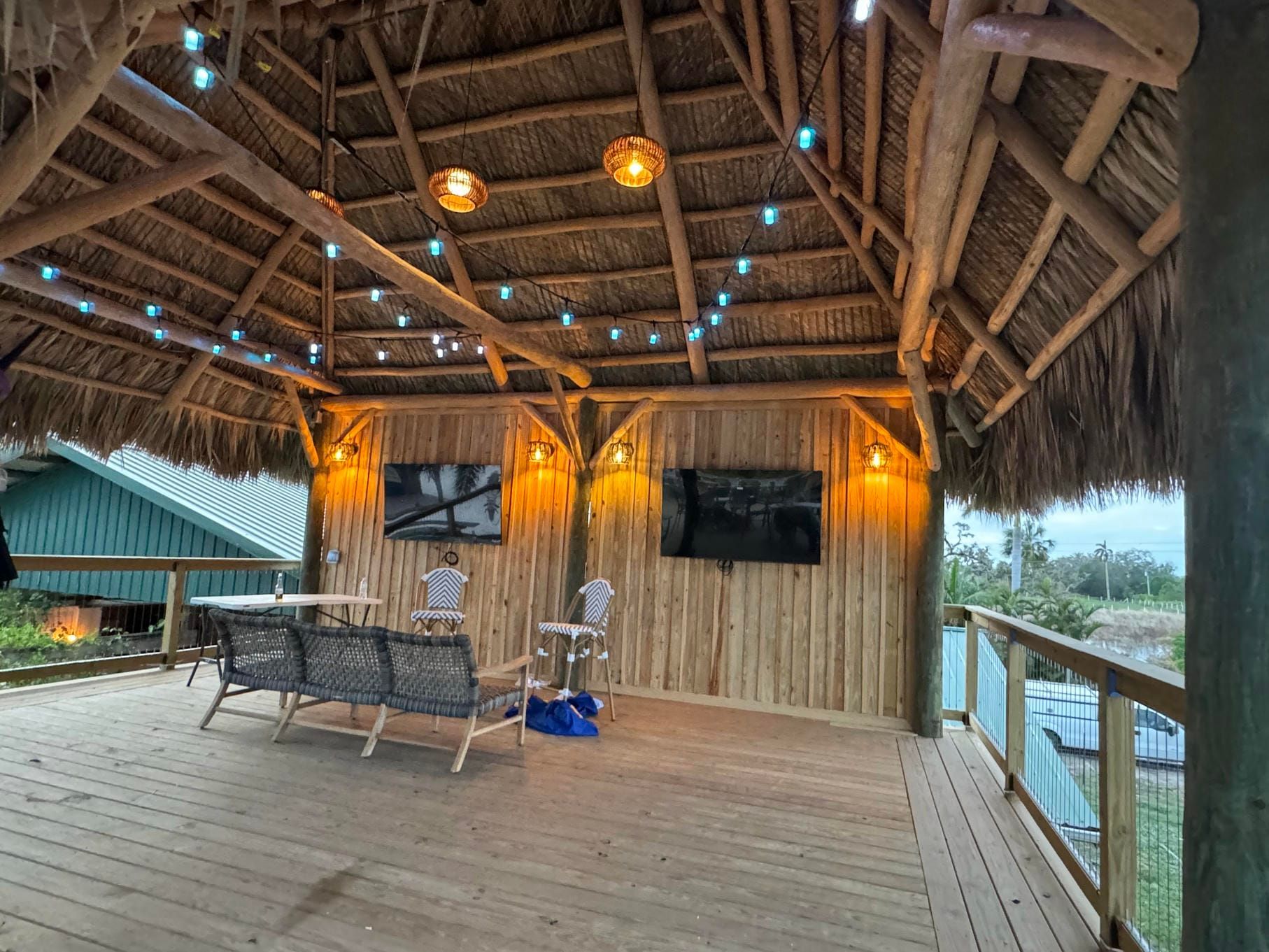 Open-air bamboo hut with wooden deck, hanging lights, and two TVs overlooking a river.
