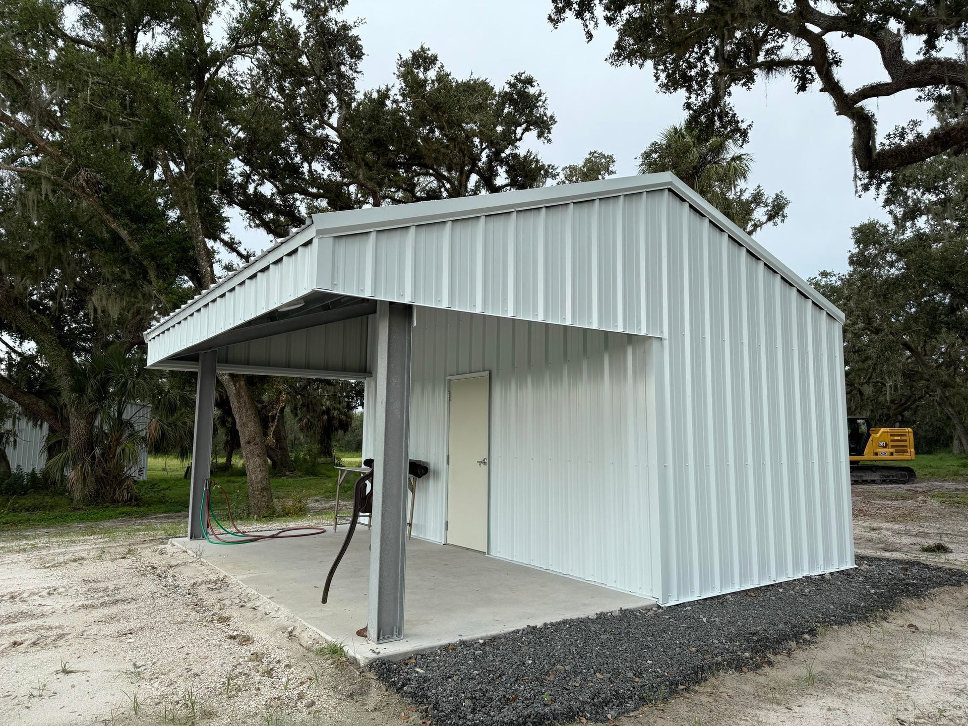 White metal shed with a covered porch and concrete floor, set on a gravel site among trees.