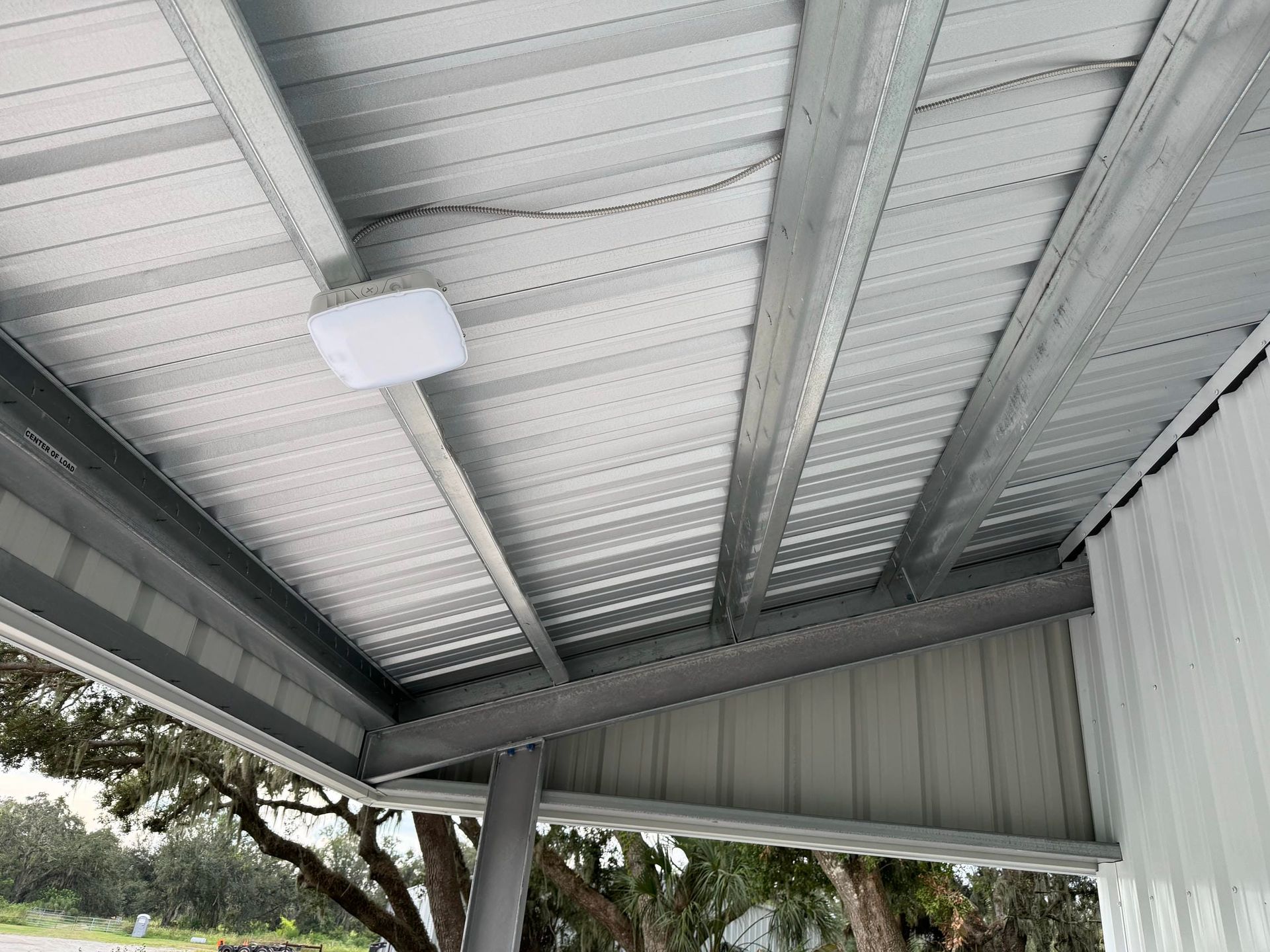 Covered patio roof with gray metal beams, white ceiling panels, and a tree visible outside.