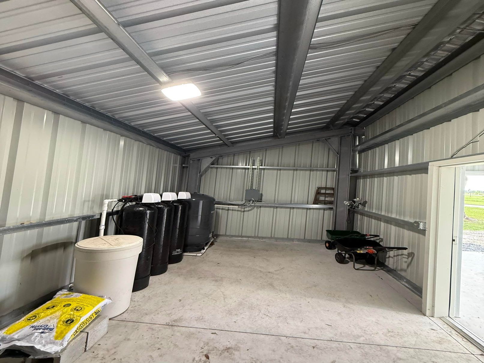 Inside a metal shed, with stacked black barrels, a white barrel, tools, and an open doorway.