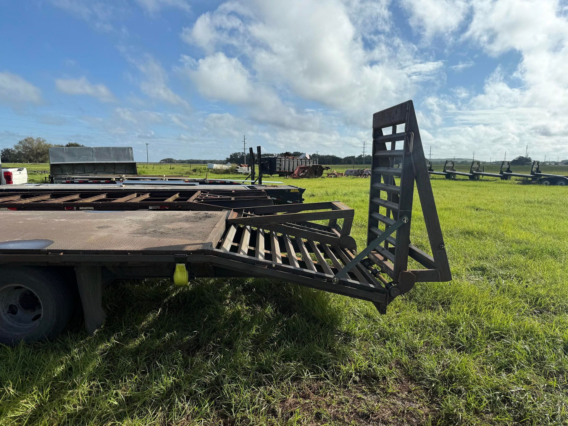Flatbed trailer with a black metal loading ramp in a grassy field under a cloudy sky