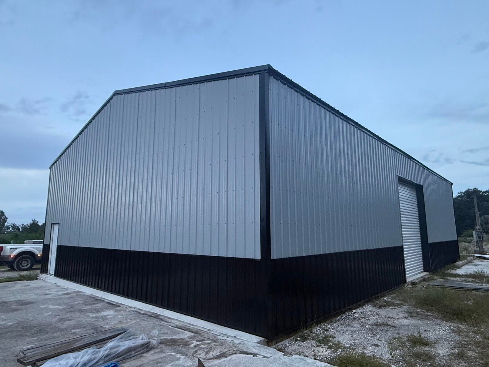 Large gray metal warehouse with black lower siding on a gravel lot under a cloudy sky