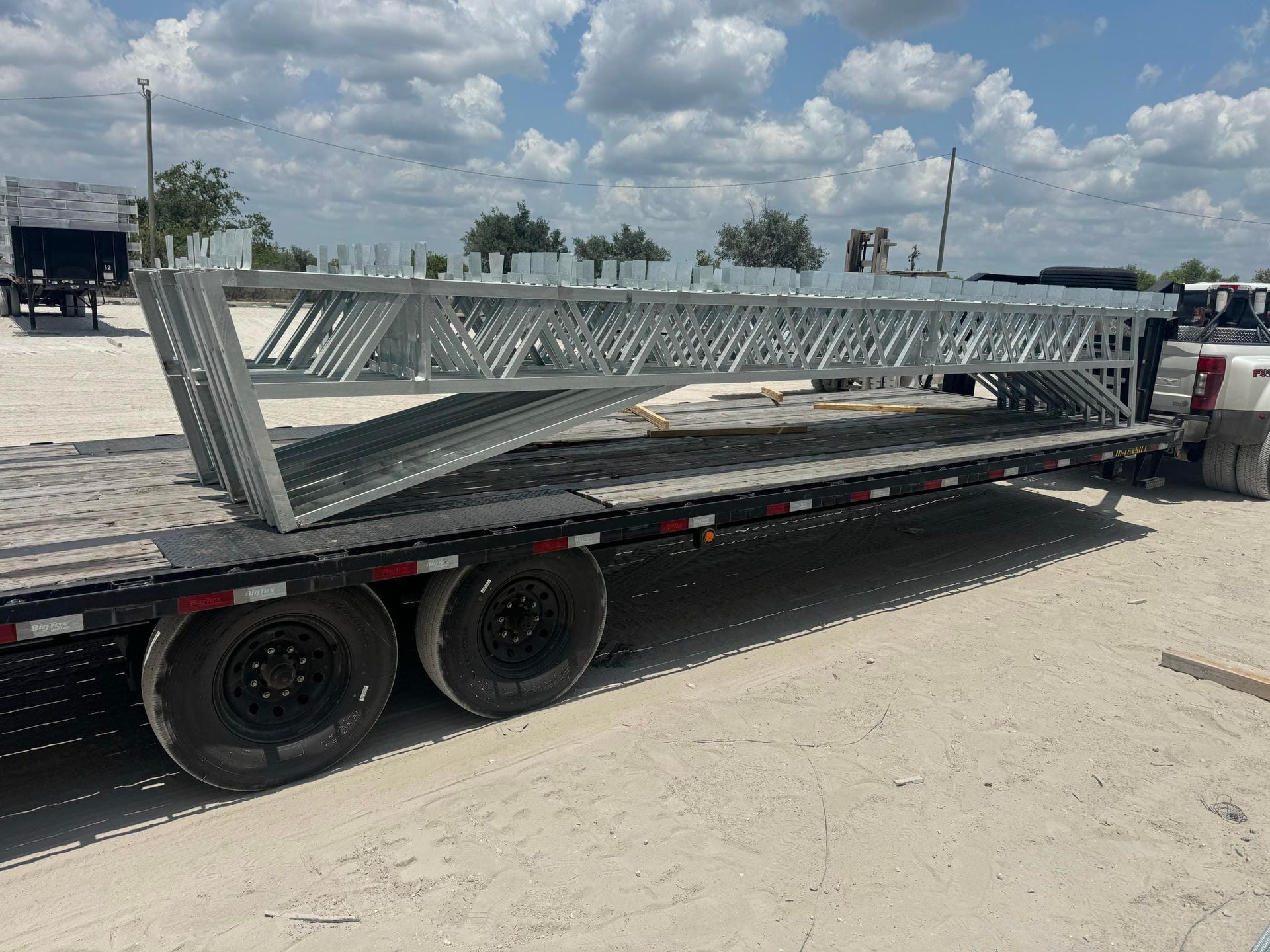 Long flatbed trailer carrying a metal truss in a parking lot under a cloudy sky