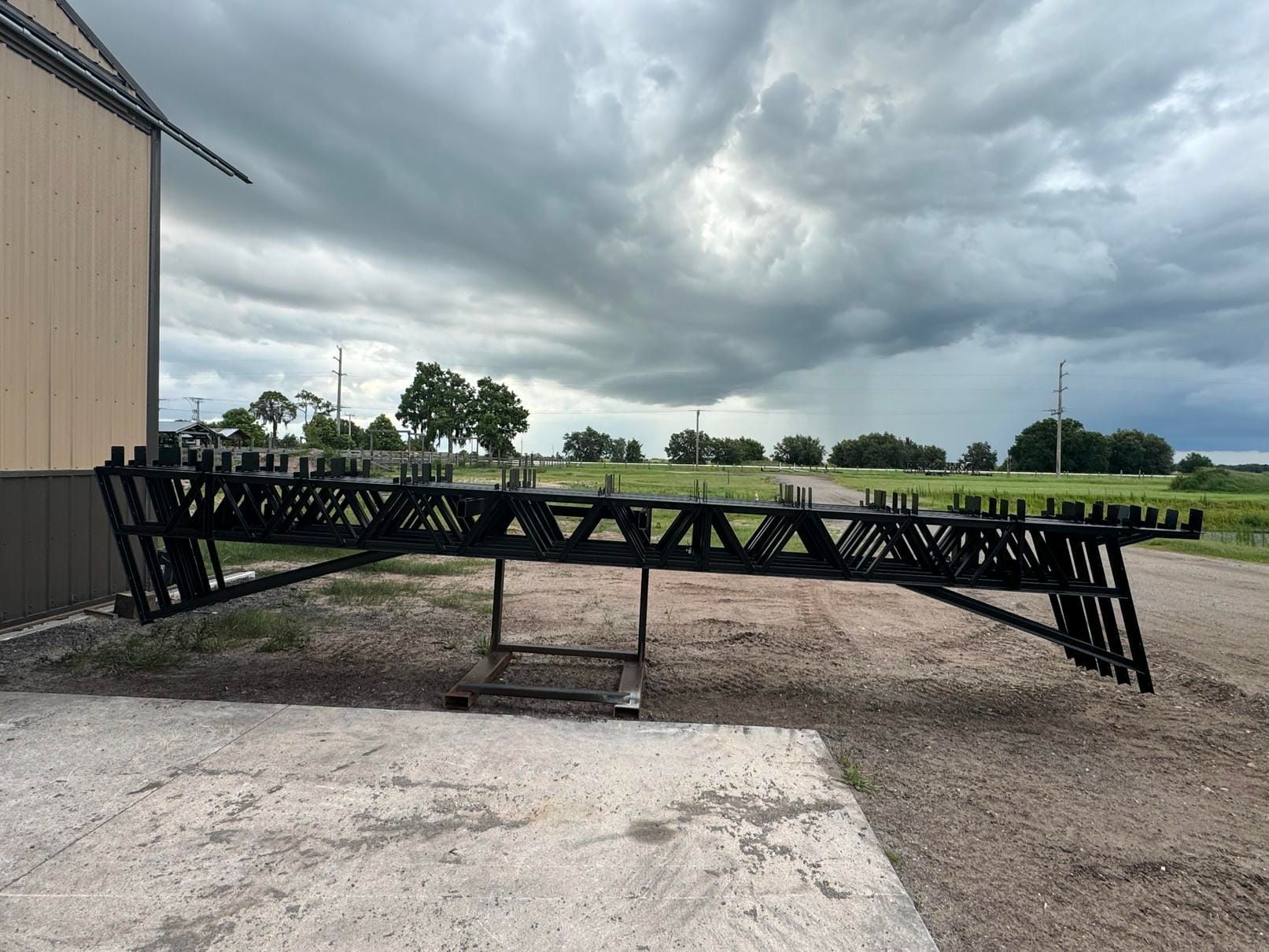 Black metal truss bridge under construction beside a building, with cloudy stormy sky overhead.