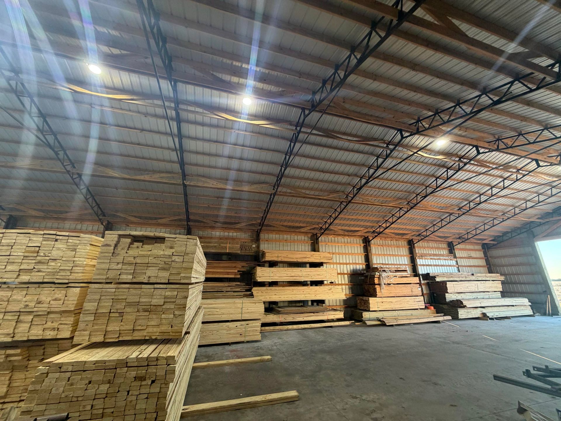 Stacks of lumber stored inside a large warehouse with a metal roof and concrete floor
