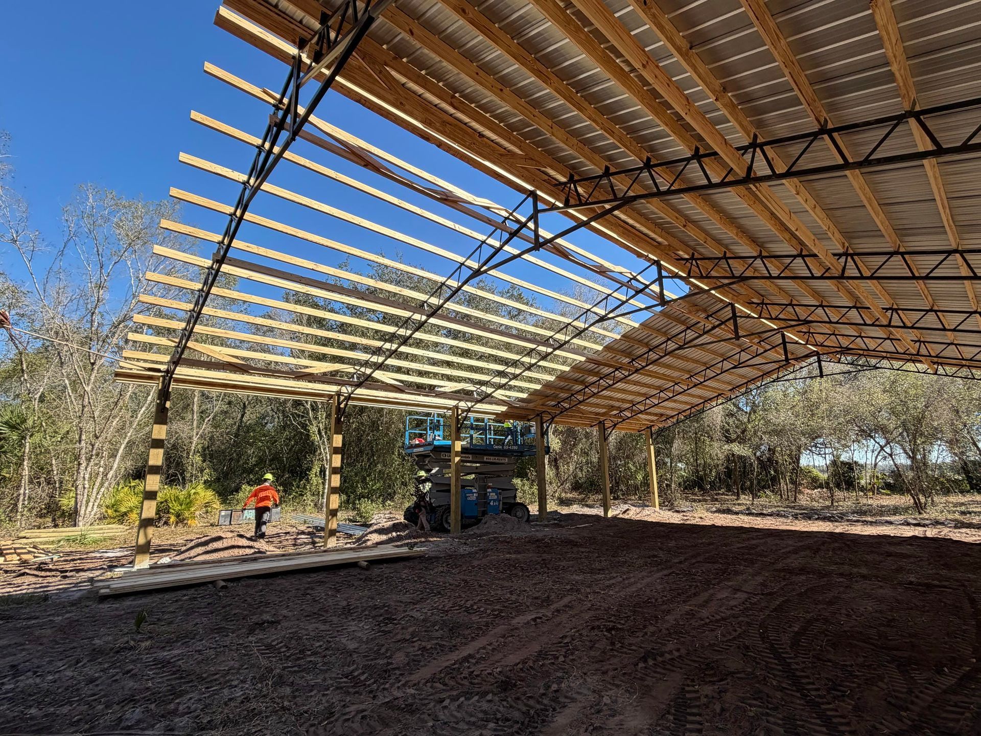 Open-sided shed under construction with wooden roof beams and a worker in orange near trees.
