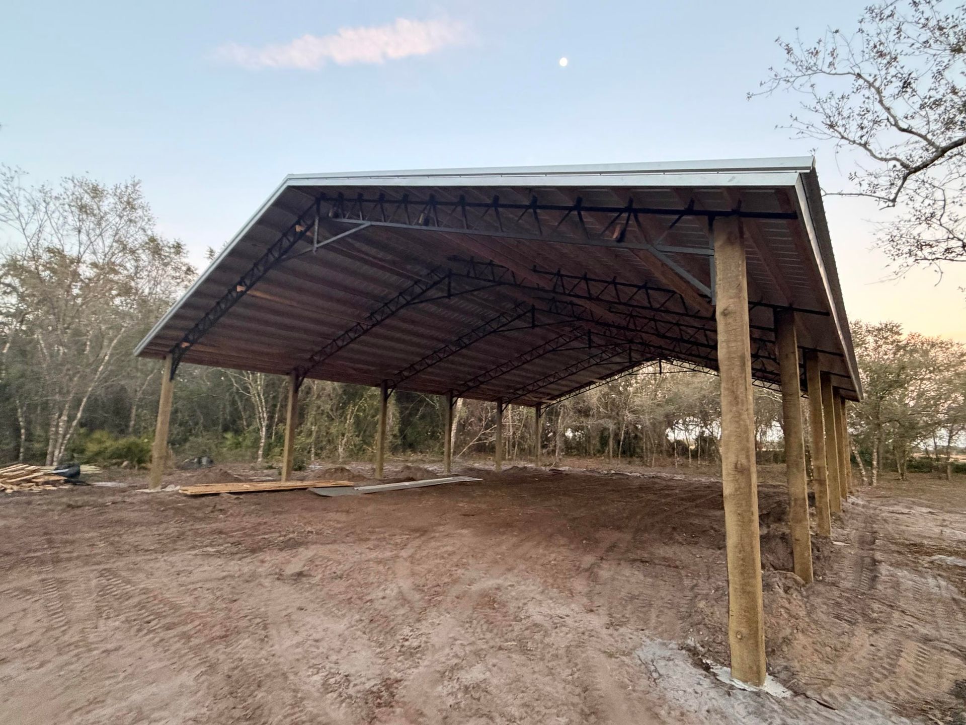 Open-sided metal-roof shelter on wooden posts in a dirt clearing with trees around it