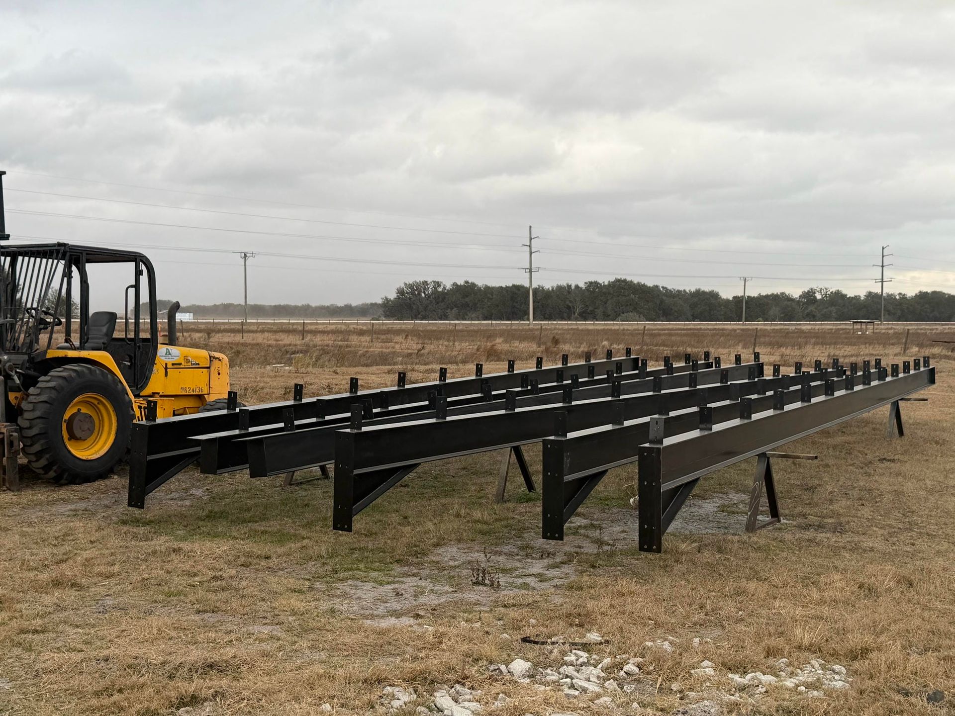 Yellow forklift beside a long black metal conveyor frame in a field under cloudy skies