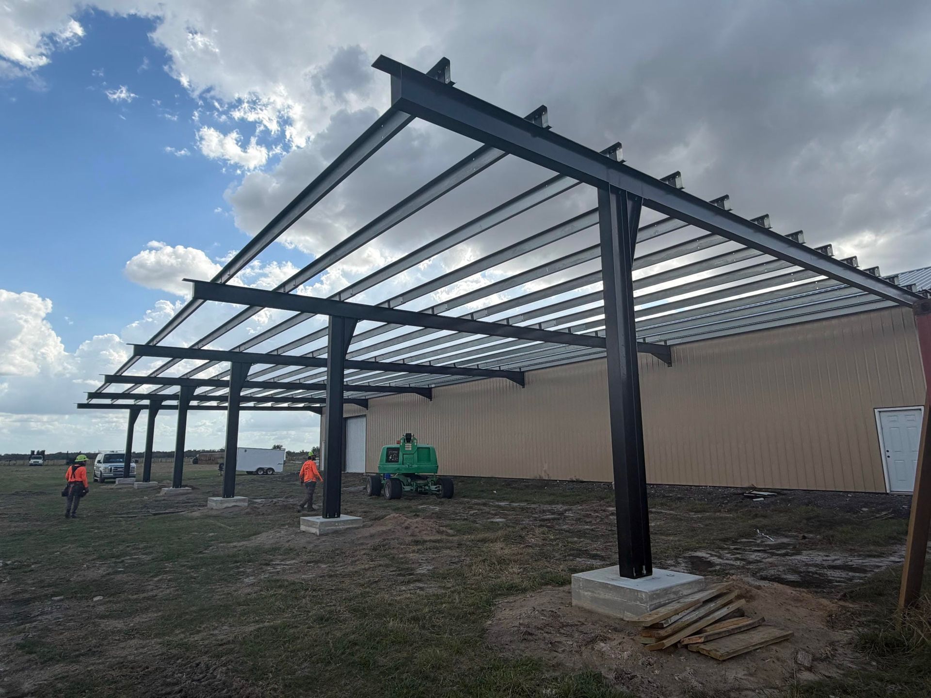 Steel canopy frame under construction beside a beige building, with workers and equipment on a grassy site.