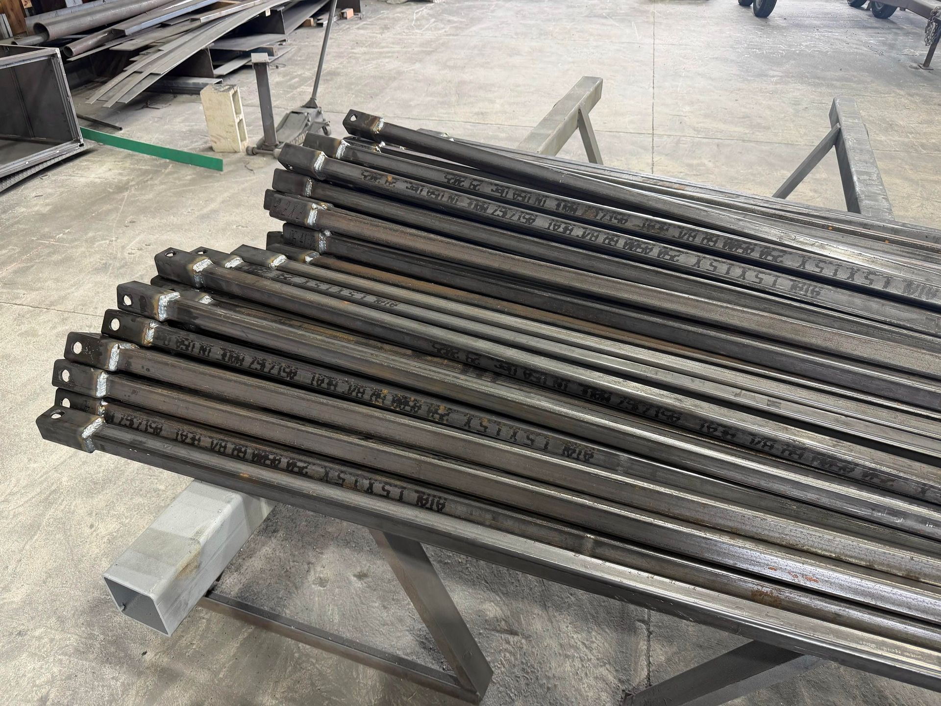 Bundle of long black metal rods stacked on a workshop floor beside a small concrete block.