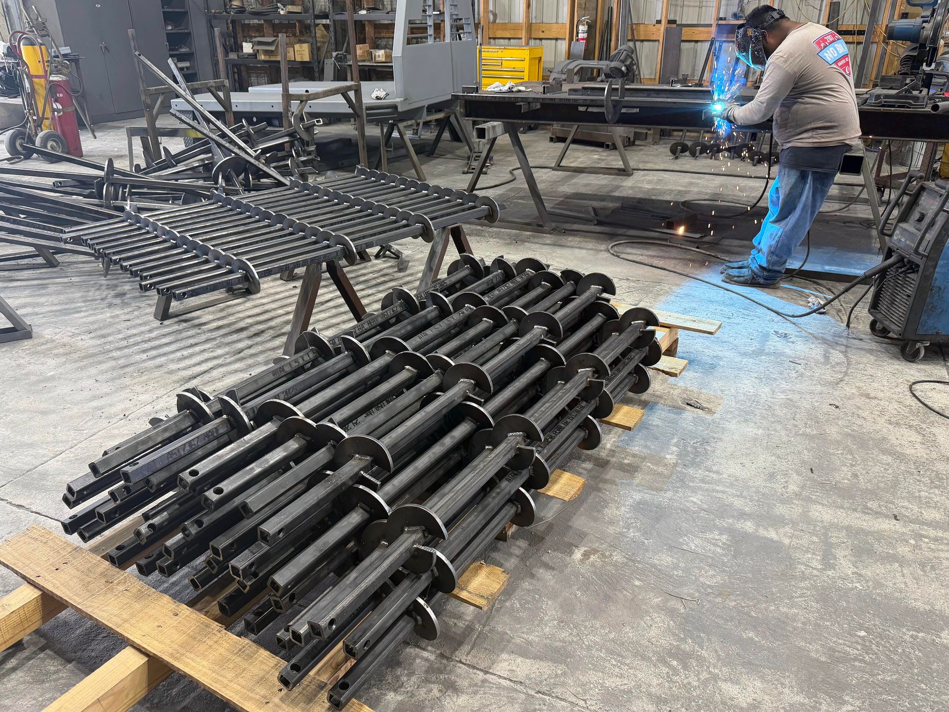 Factory worker welding black metal frames beside stacked parts in an industrial workshop.