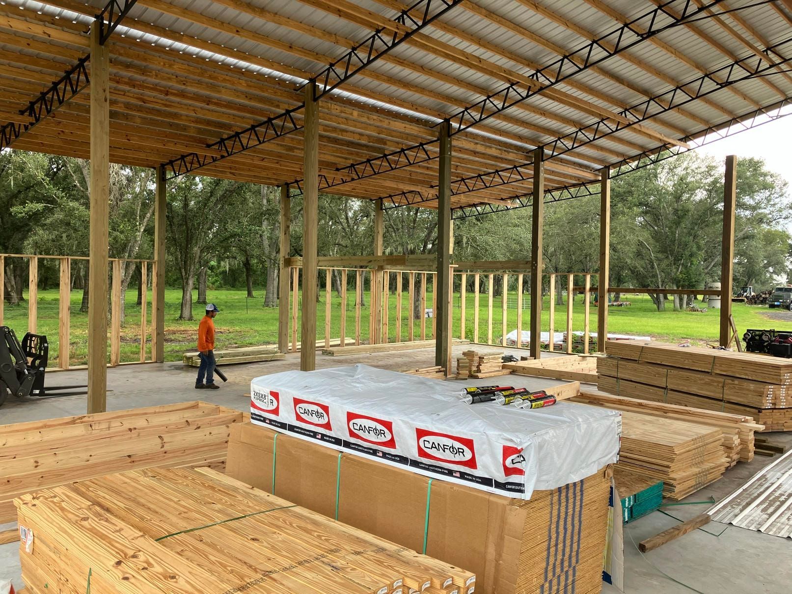 Construction site with steel roof frame, stacked lumber, and a worker in orange on a concrete slab