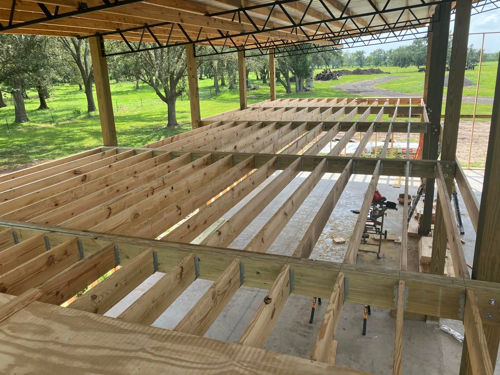 Wooden roof framing under construction beneath a metal canopy in a grassy outdoor area