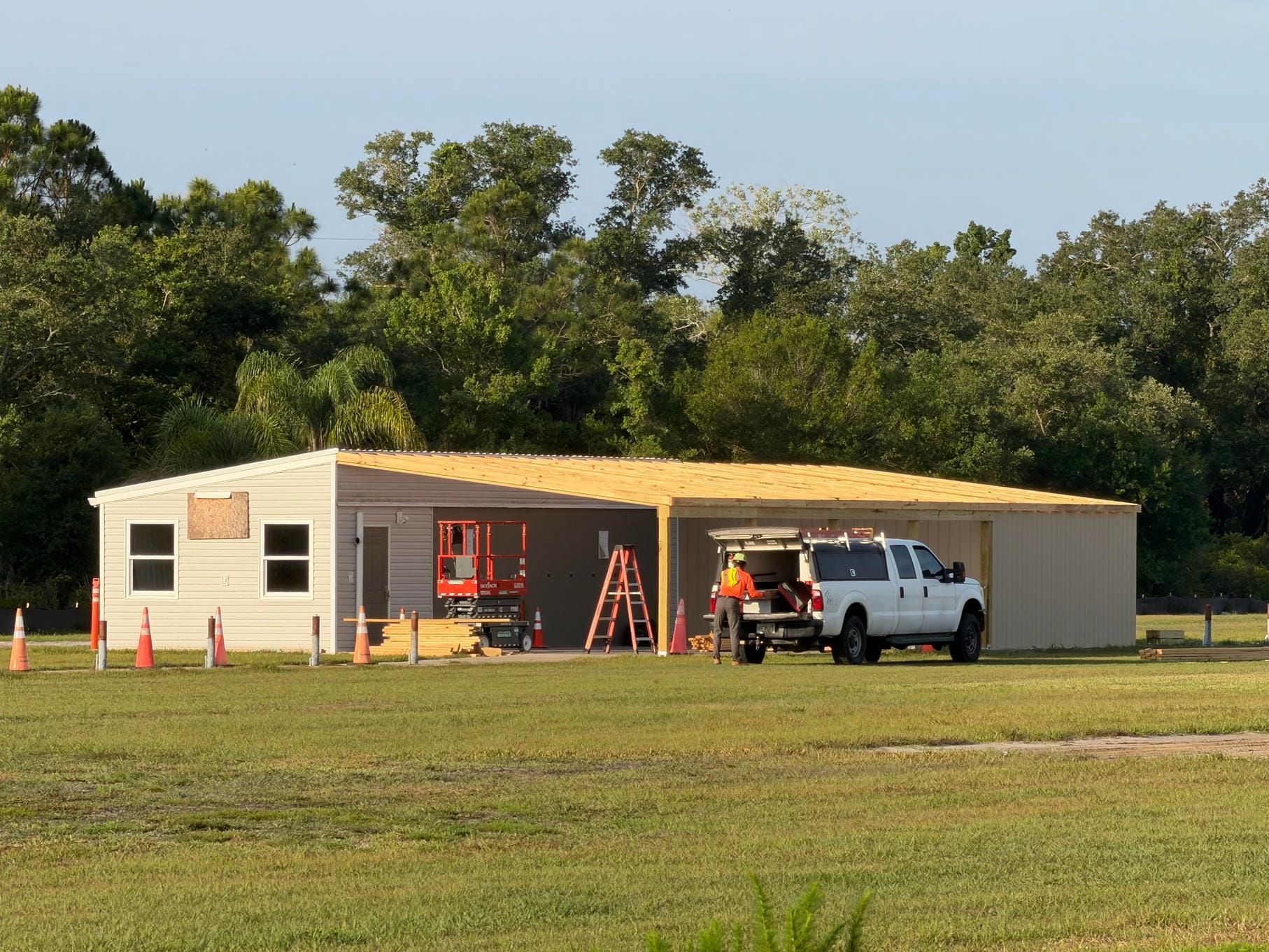 Portable building and white pickup truck on a grassy field, with ladders and orange cones outside.