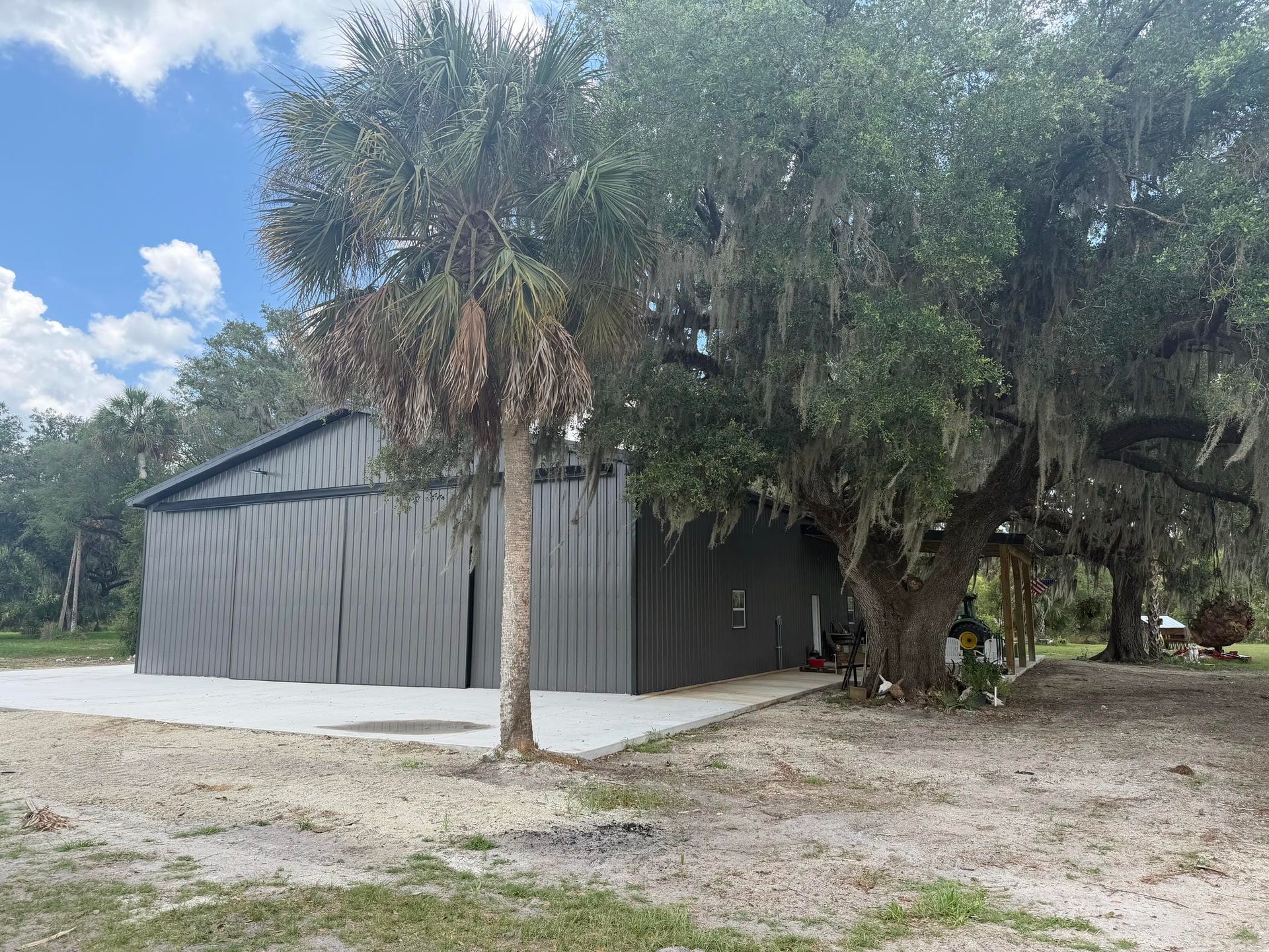 Gray metal barn beside palm trees on a gravel lot under a partly cloudy sky