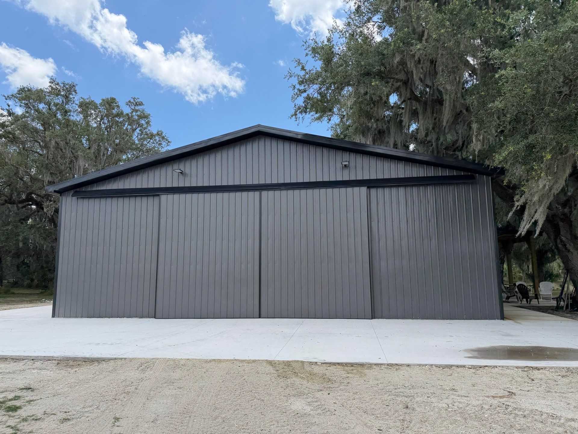 Large dark gray metal building with a peaked roof on a white gravel lot under a blue sky.