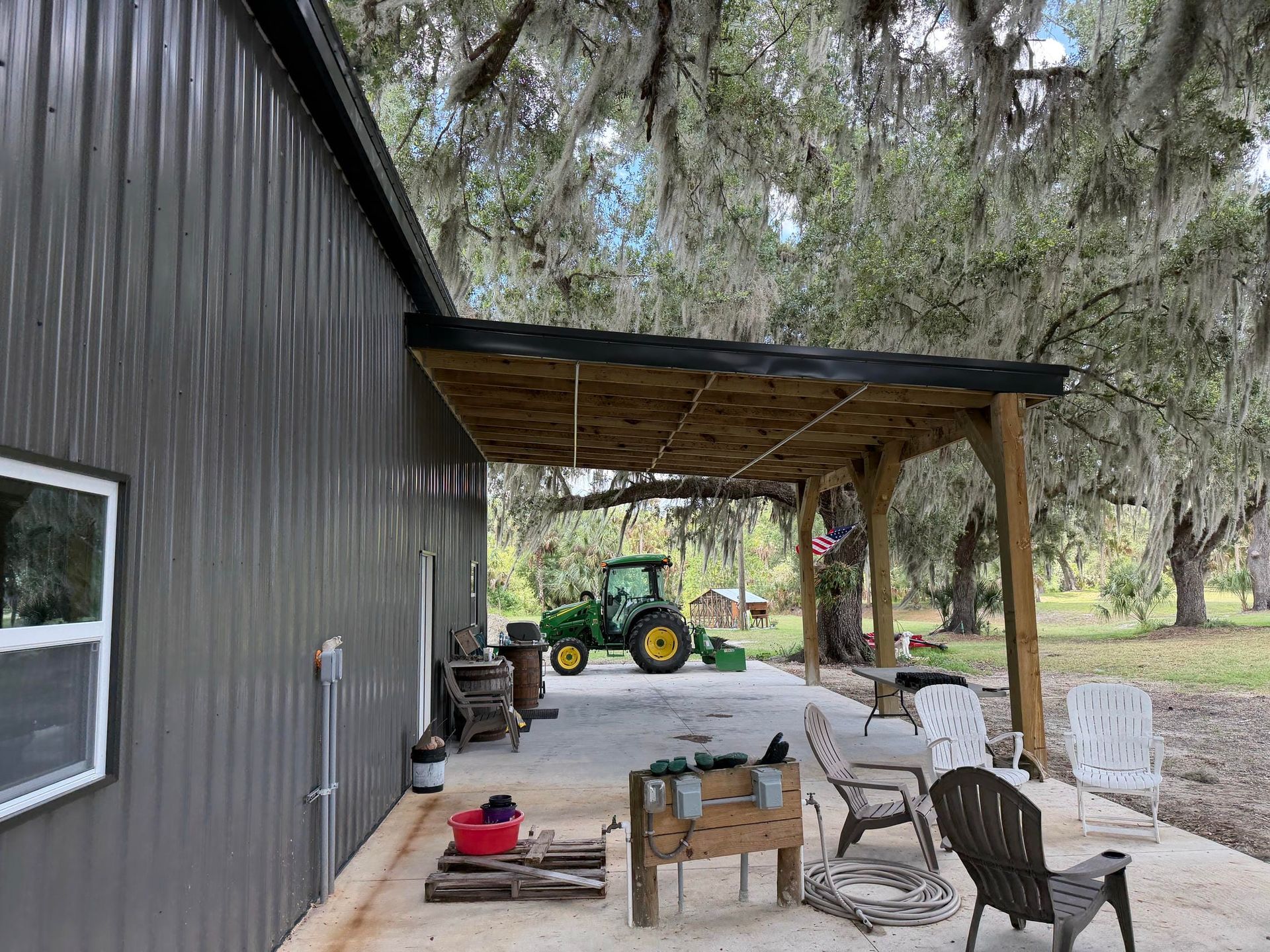 Covered porch with chairs and table beside a dark metal house, overlooking a yard with a green tractor.