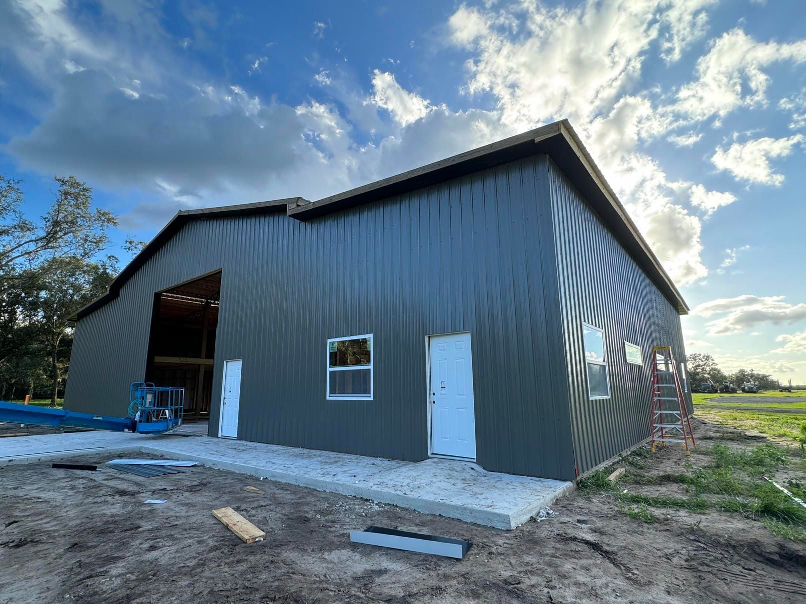 Partially built metal building with corrugated siding, exposed framing, and an orange lift under a blue sky