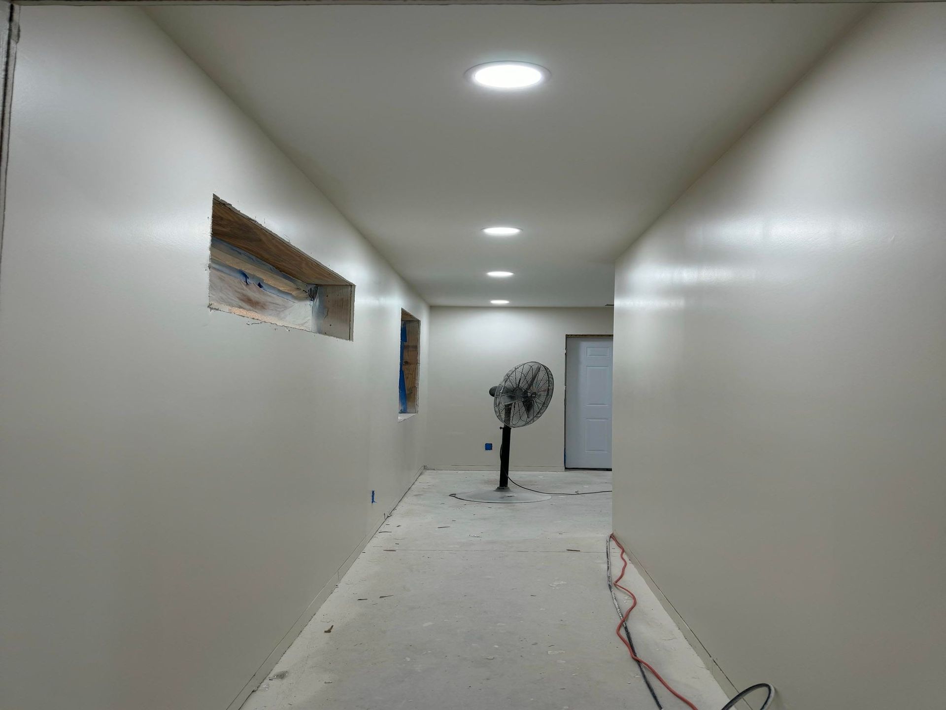 Narrow white hallway under construction with recessed lights and a fan at the far end.