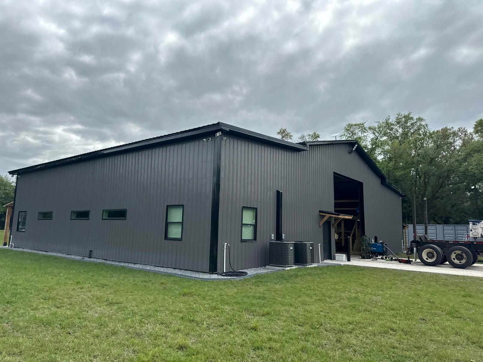 Gray metal workshop building with open garage bay, small windows, and a tractor parked beside it under cloudy skies