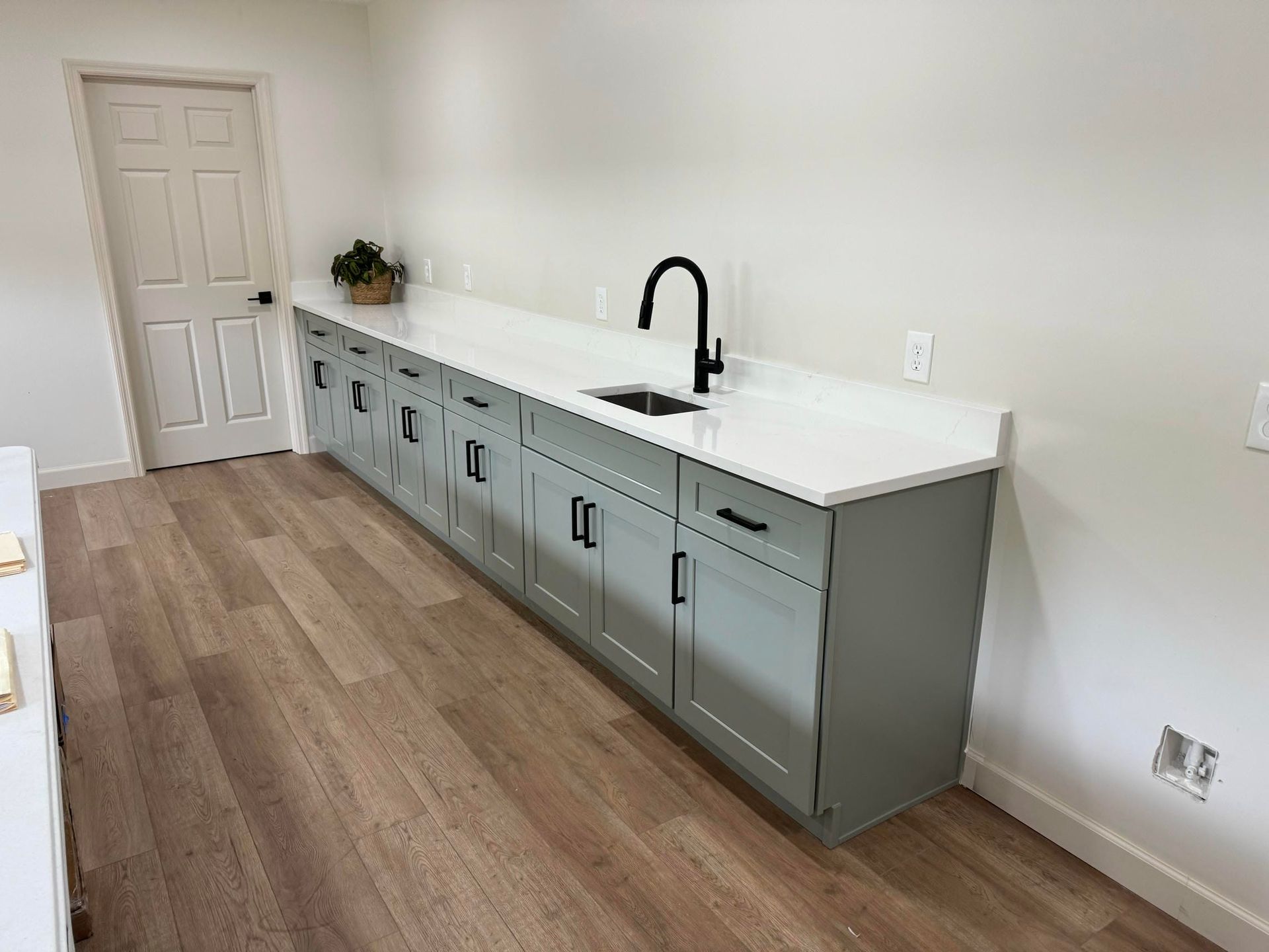 Long narrow room with gray cabinets, white countertop, black faucet, and wood-look flooring.