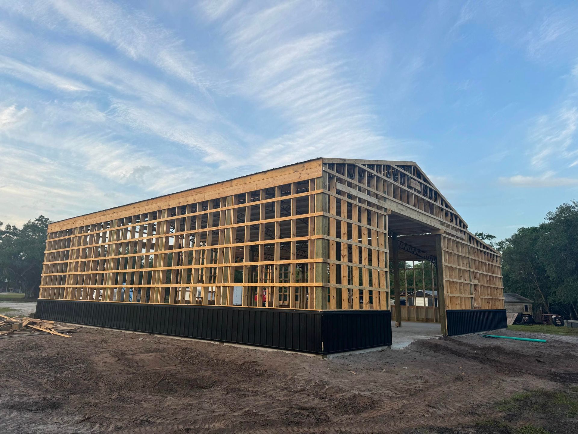 Large wooden building under construction on a dirt lot at sunset, with trees and a blue sky.