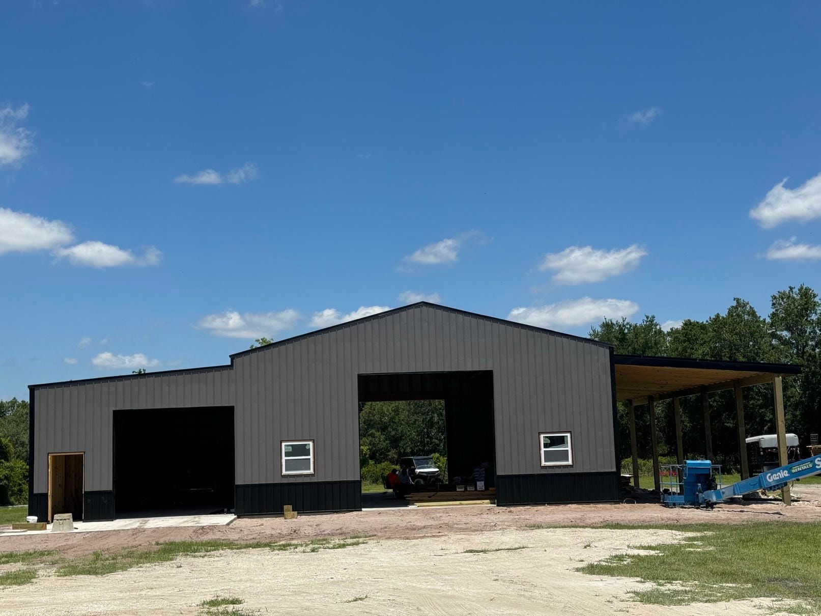 Gray metal barn under construction on a dirt lot, with open doors and blue sky.