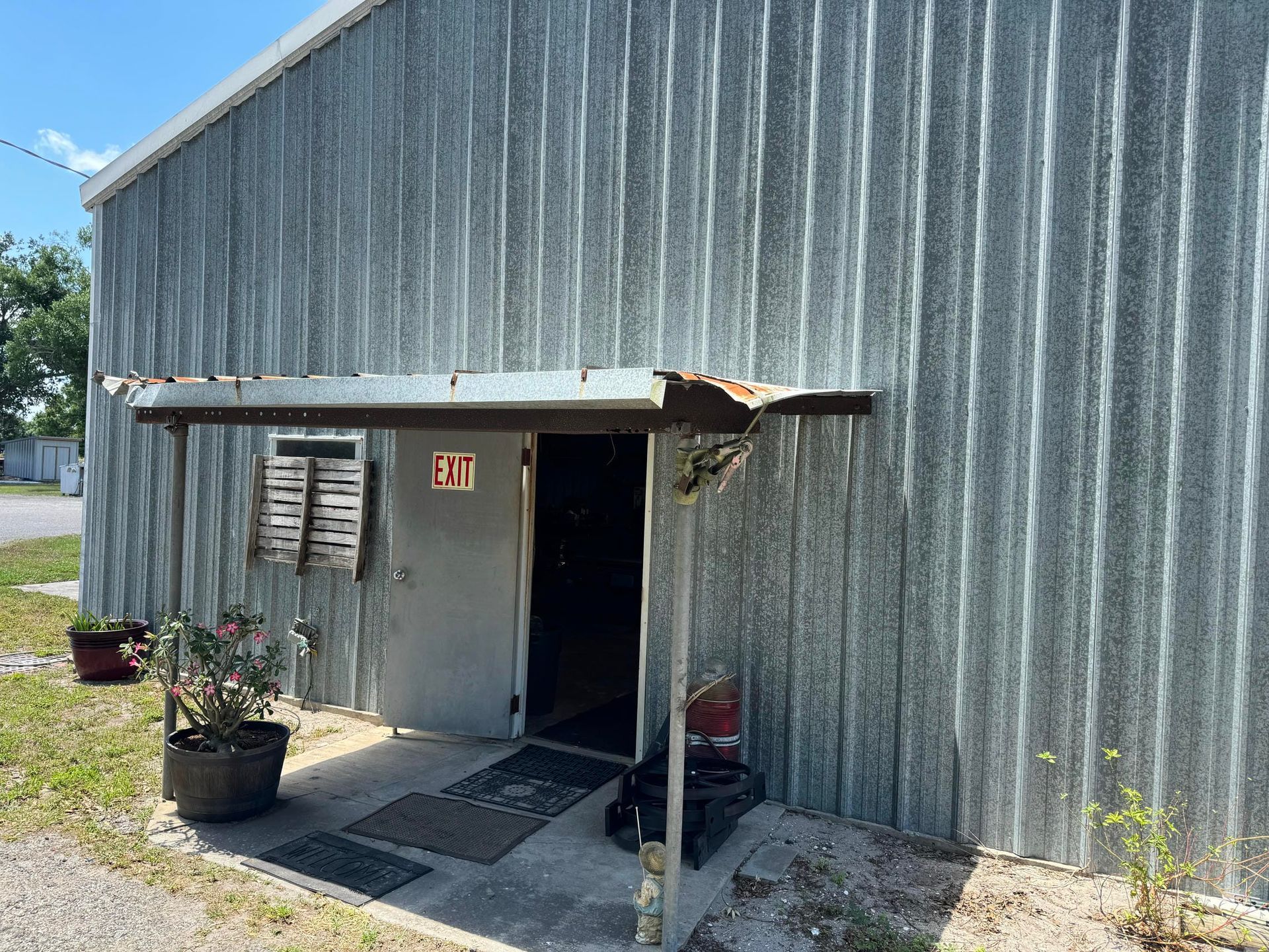 Gray metal building entrance with a small awning, potted plants, and a side door in a sunny outdoor setting