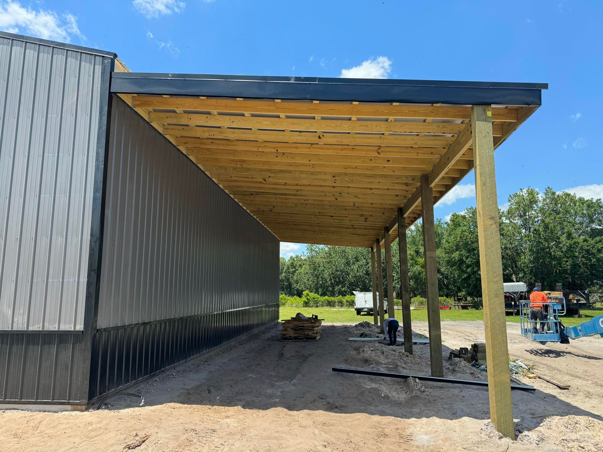 Partially built wooden roof canopy attached to a black building on a gravel lot under a blue sky