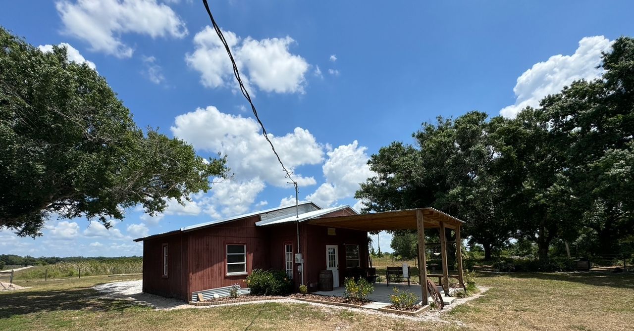 Small red building with a porch under a blue sky, surrounded by trees and a field.